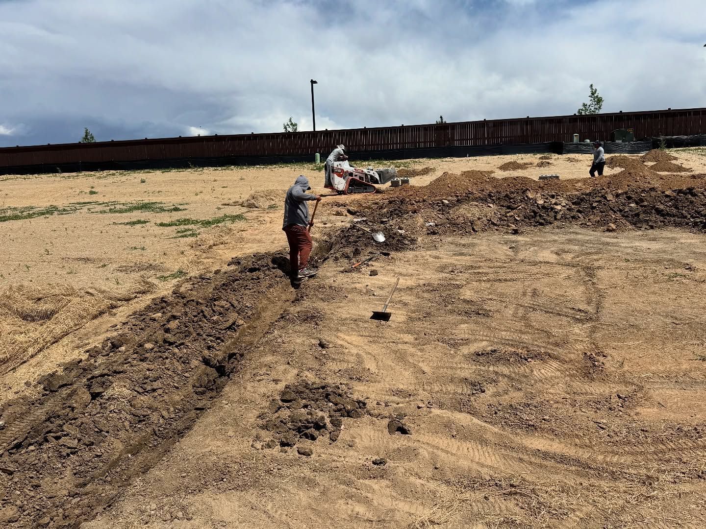 Two people dig a trench in a large, sandy, open construction site under a partly cloudy sky.