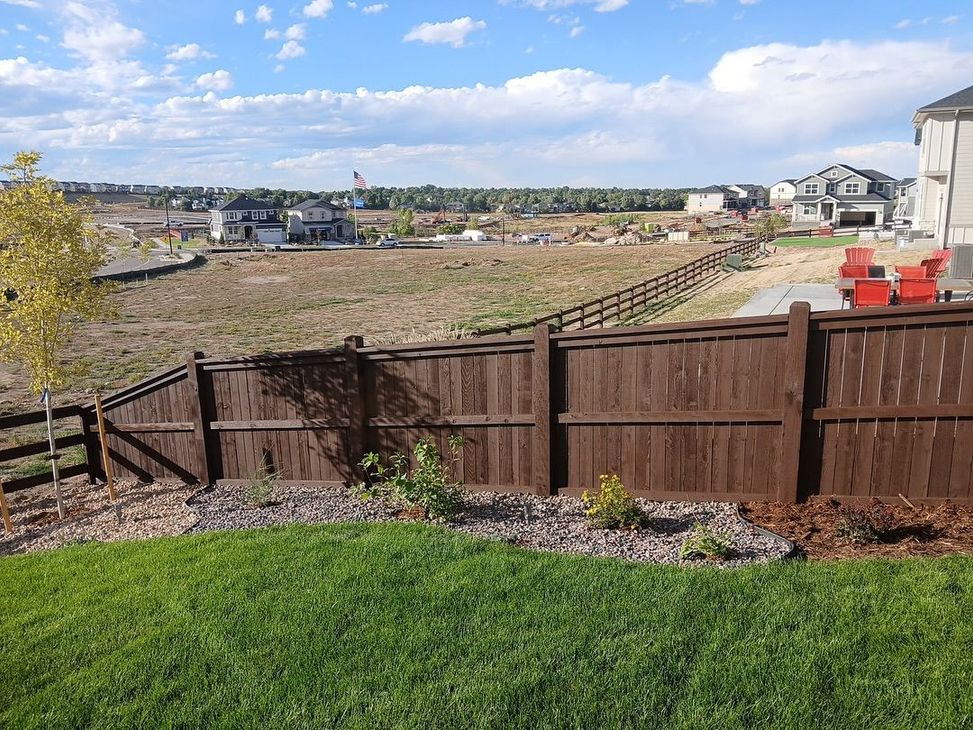 A brown wooden fence runs across a sloped grassy yard with landscaping, overlooking a field and suburban houses.