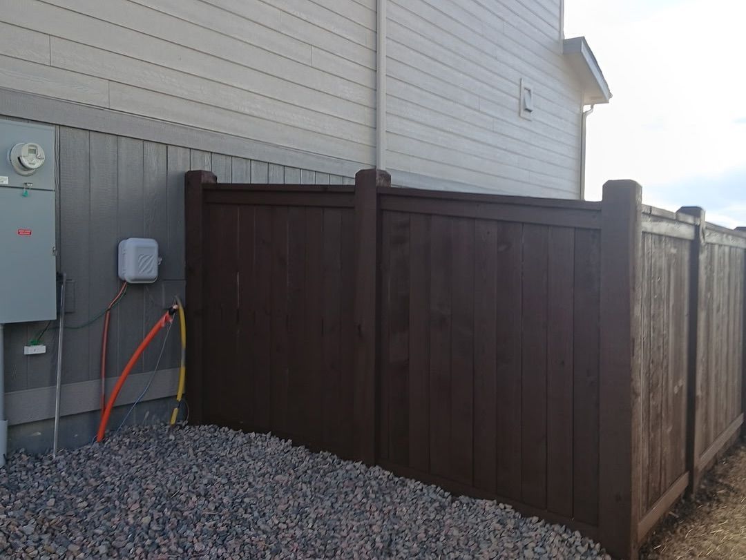 A dark brown wooden fence sits adjacent to the side of a light-colored house exterior with exposed utility components.