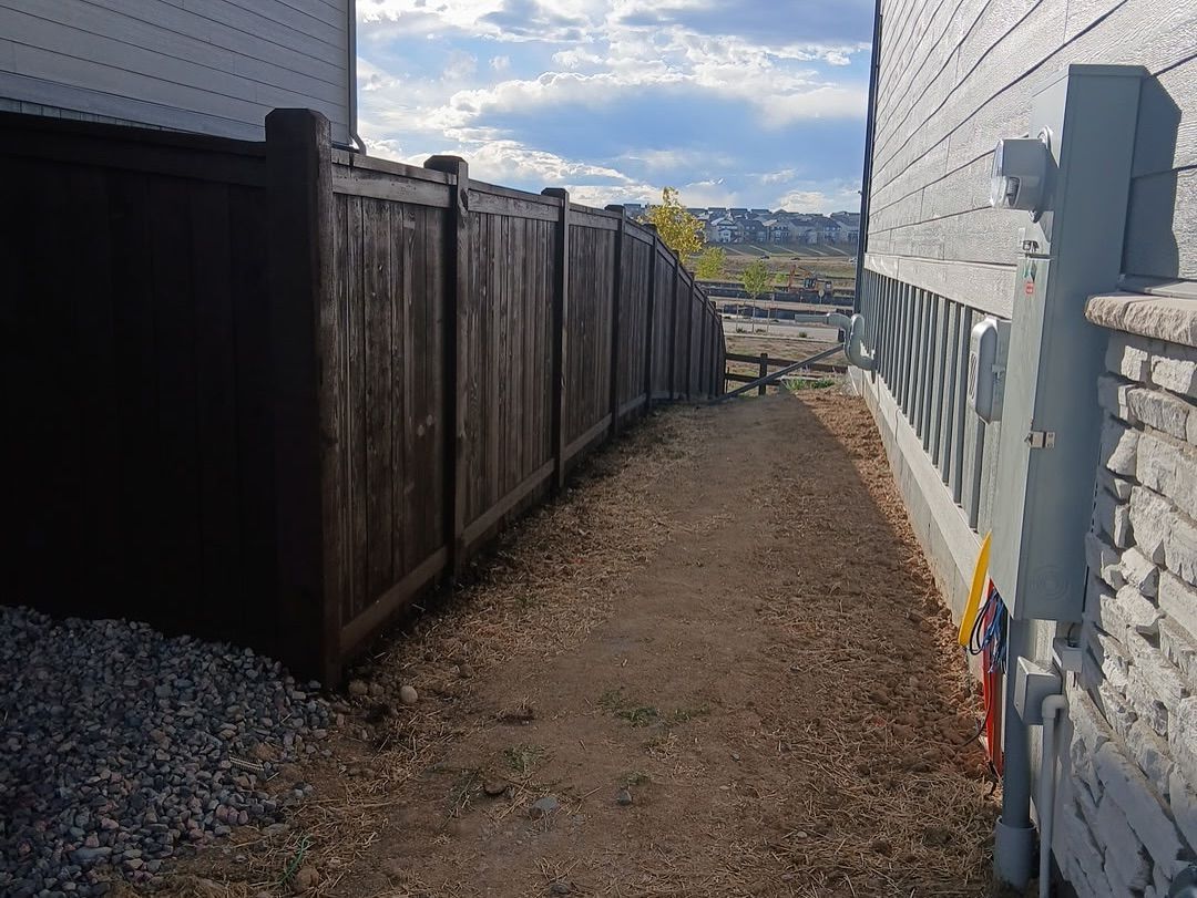 A narrow dirt path between a wooden fence and a house, leading toward an open area under a cloudy sky.