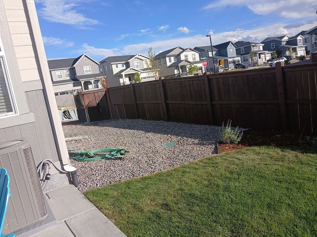 A backyard scene featuring a stone-covered area adjacent to a grassy lawn, enclosed by a dark brown wooden privacy fence.