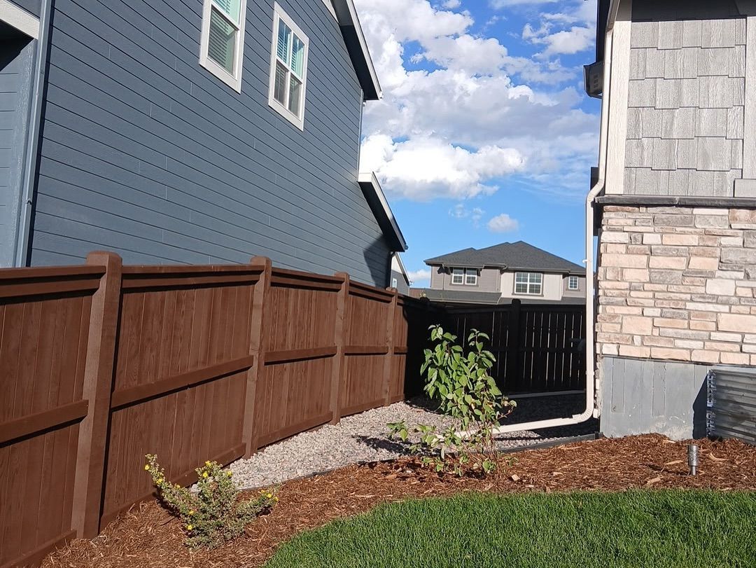 A brown wooden fence separates two suburban houses with gravel landscaping and small plants in the yard.