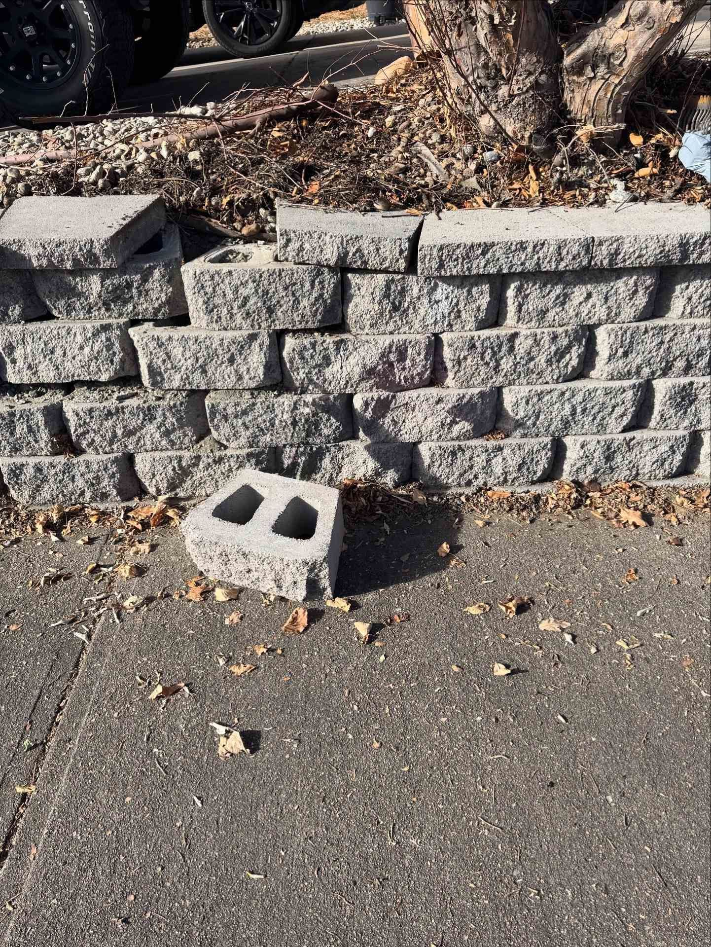 A broken concrete retaining wall on a sidewalk with one detached, hollow block sitting on the ground in front of it.