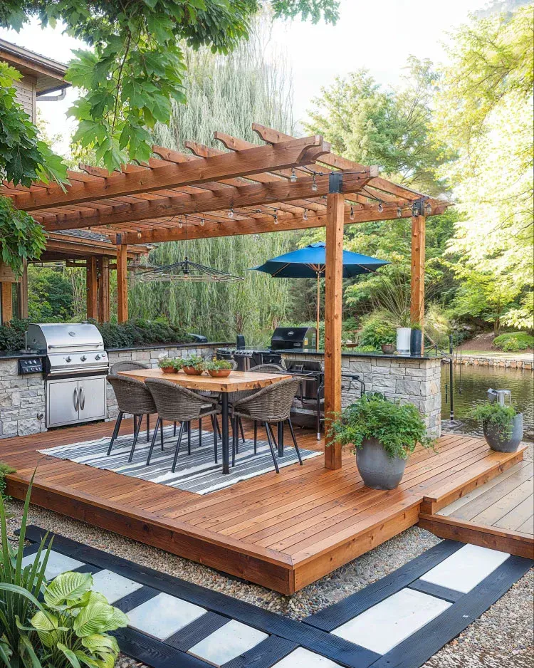 Outdoor stone patio with a built-in kitchen, stainless steel refrigerator, wooden cabinets, and trees in the background.