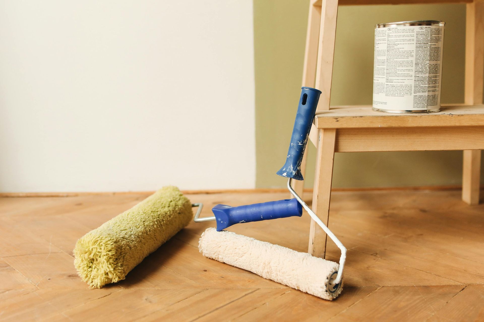 A paint roller, a spare roller cover, and a paint can on a step stool sit on a wooden floor in front of a painted wall.