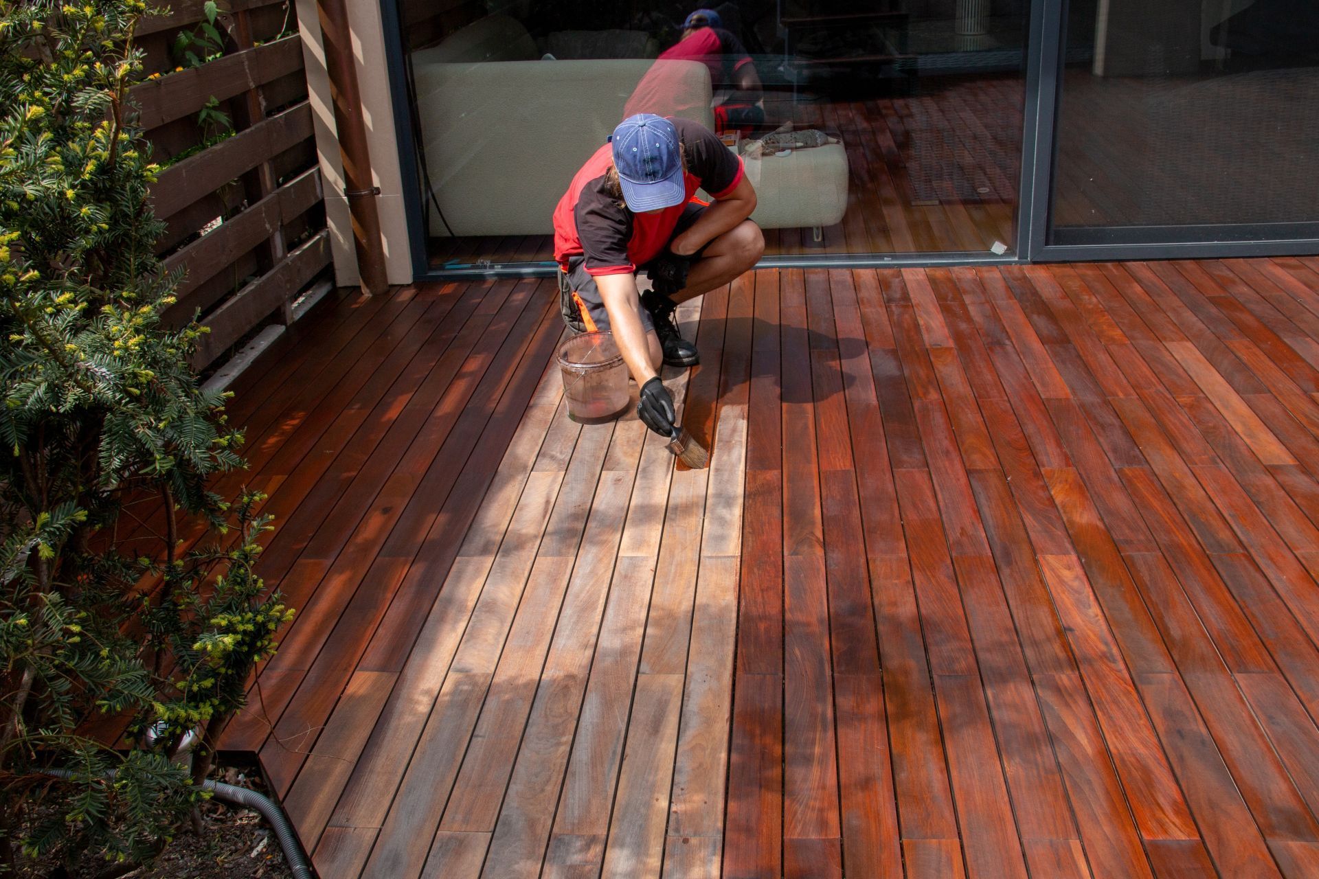 A person in a red shirt and blue cap kneeling on a wooden deck, applying stain to a lighter section of wood.