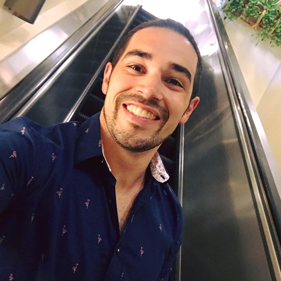 A smiling person with a beard wears a blue, flamingo-patterned button-down shirt while standing near an escalator.