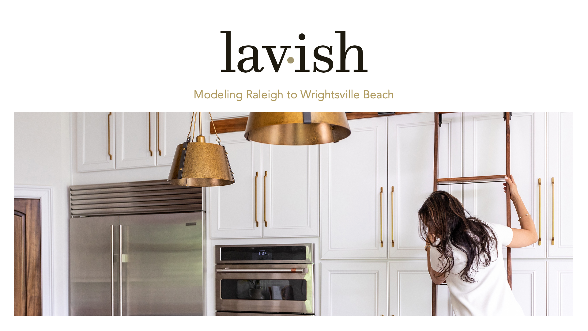A person climbs a library-style ladder in a modern white kitchen with gold hardware and hanging gold pendant lights.