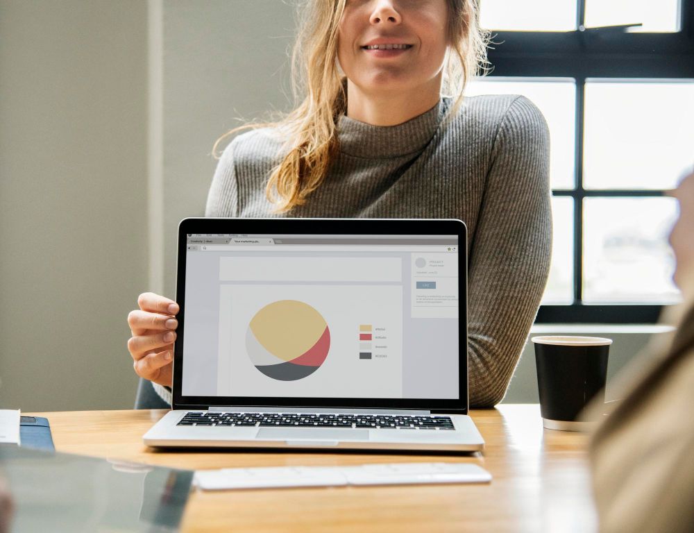 a woman is sitting at a table holding a laptop computer with a pie chart on the screen .