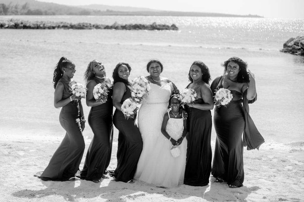 Bride with bridesmaids on a beach, smiling. All wear long gowns, hold bouquets. Black and white photo.