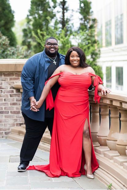 Couple, smiling, holding hands. Woman in red gown, man in blue shirt. Outdoor setting, stone wall.