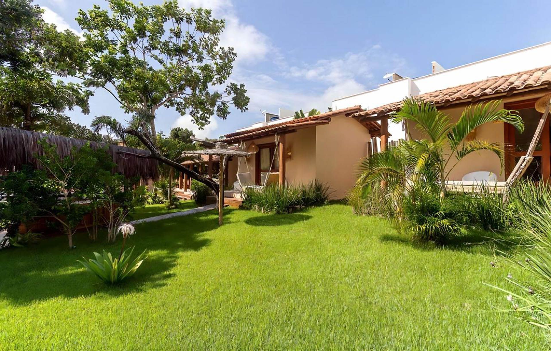 Quintal ensolarado com gramado verde, árvores e uma casa com telhado de telha e pátio com vista para o jardim.