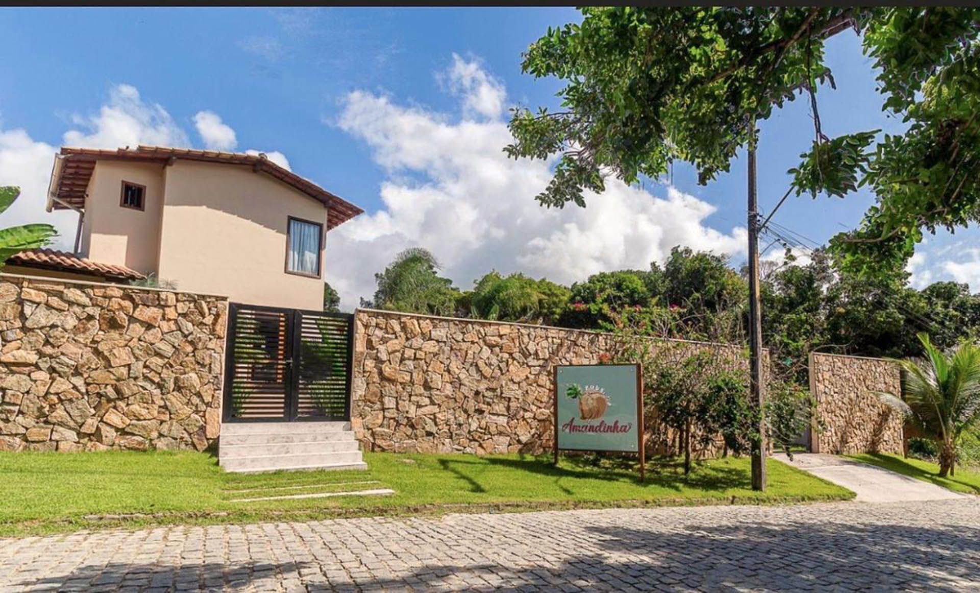 Entrada com muro de pedra para uma casa moderna com portão de vidro, gramado verde e céu azul com nuvens.