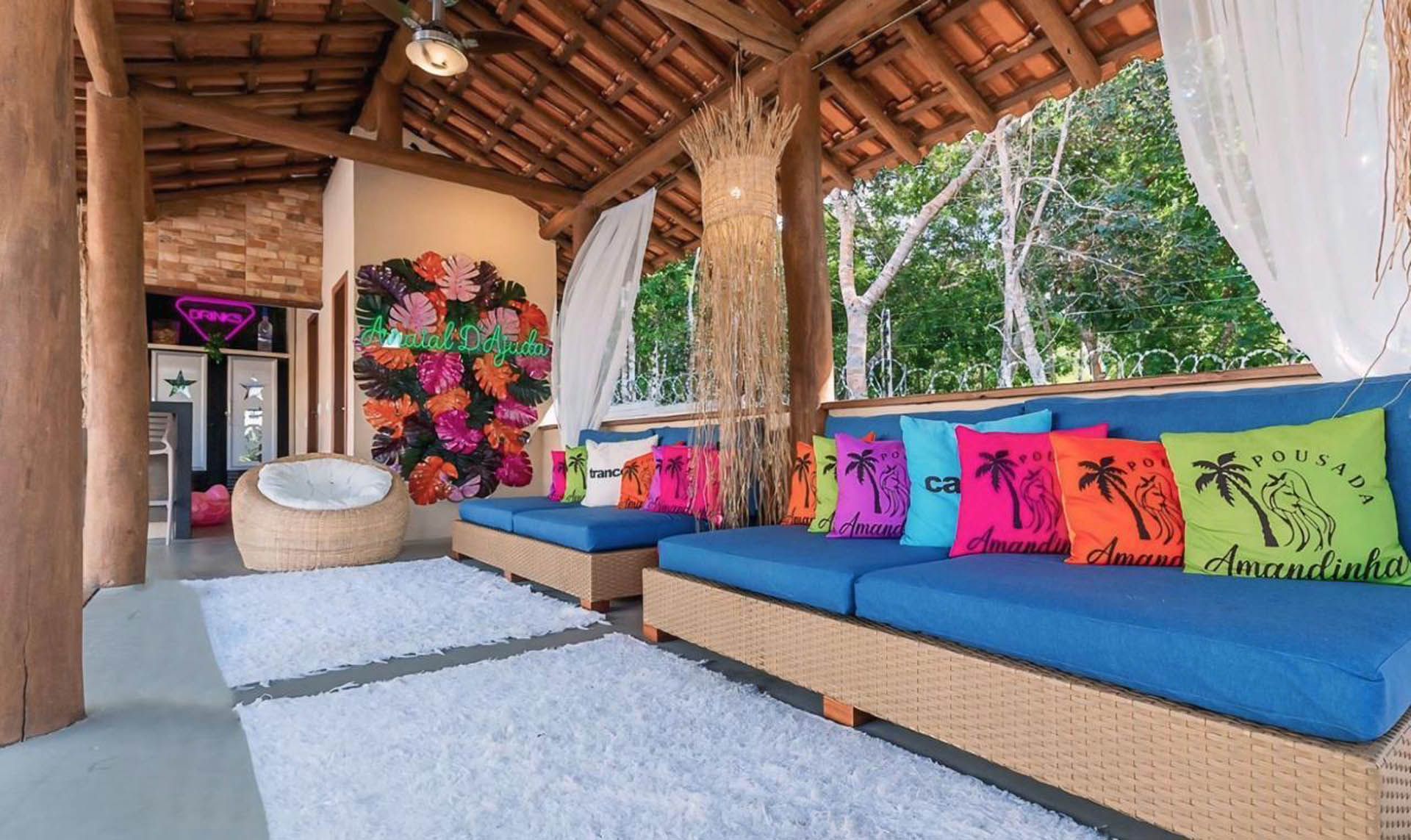 Lounge ao ar livre com assentos azuis e bege, almofadas coloridas e teto de palha em um ambiente tropical.