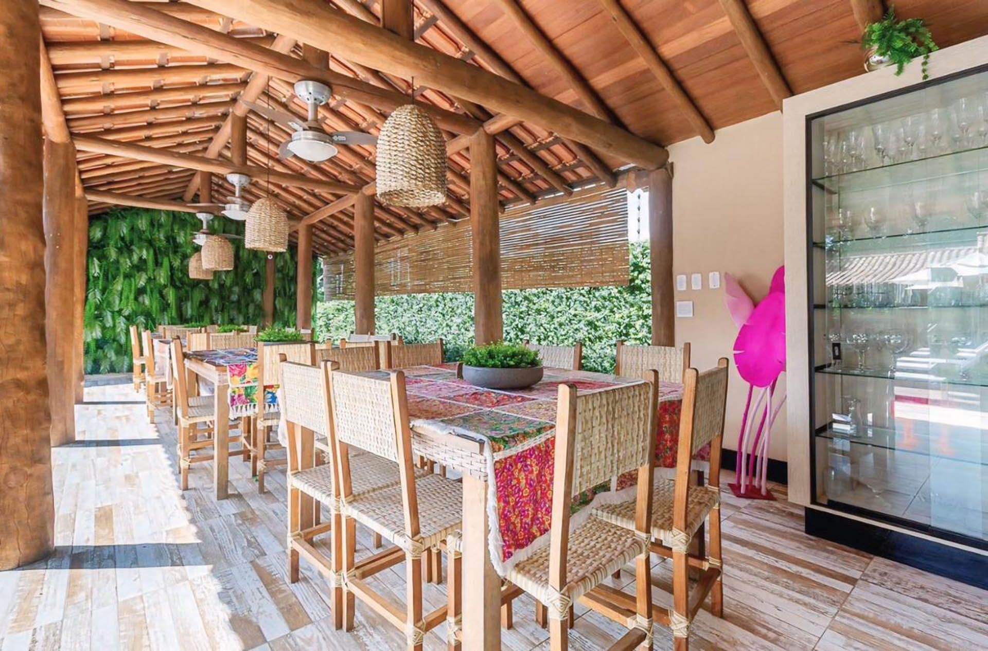 Sala de jantar ao ar livre com mesa de madeira, cadeiras de vime, luminárias pendentes e geladeira com porta de vidro à direita.