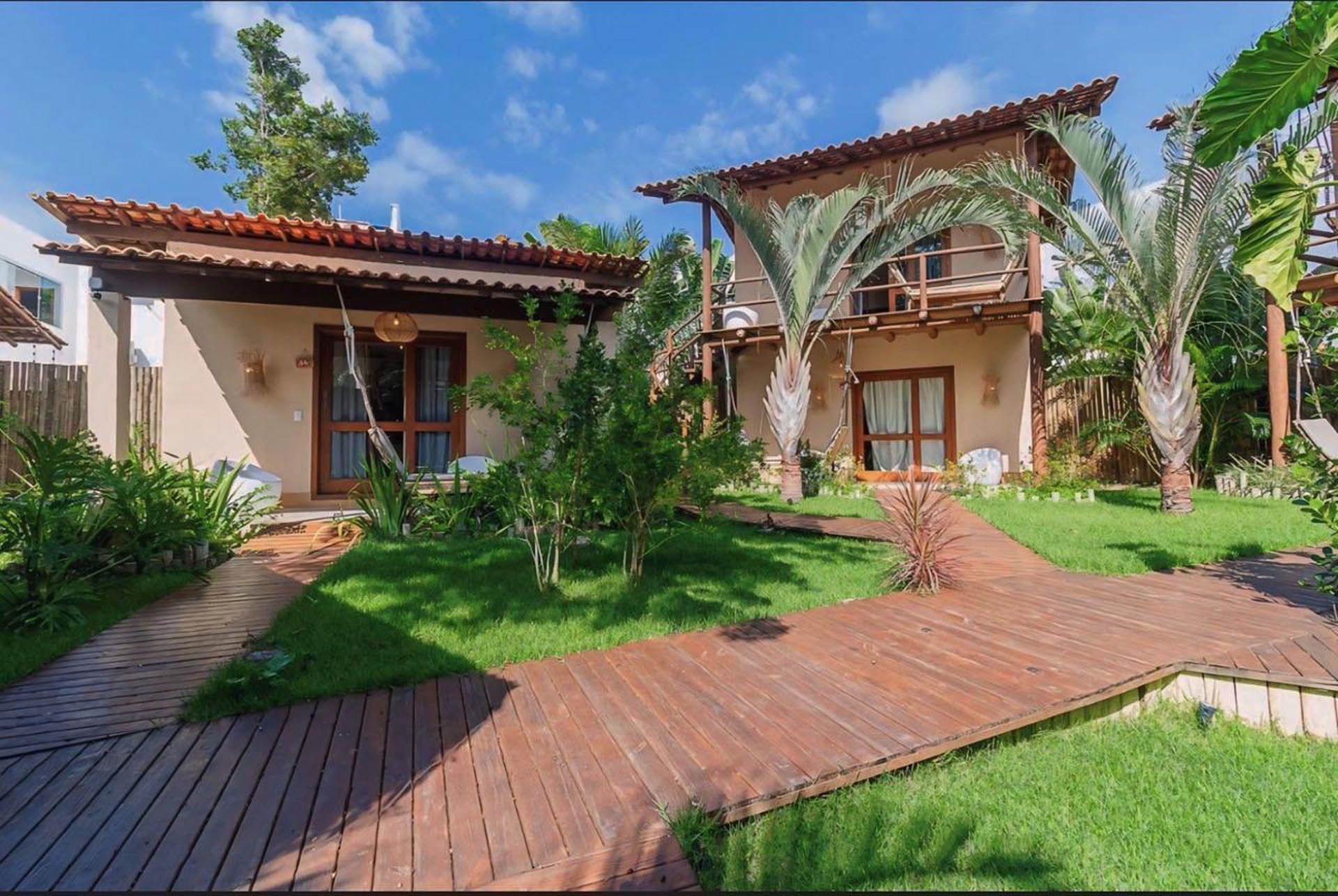 Jardim tropical com duas pequenas casas de campo, passarela de madeira e céu azul.