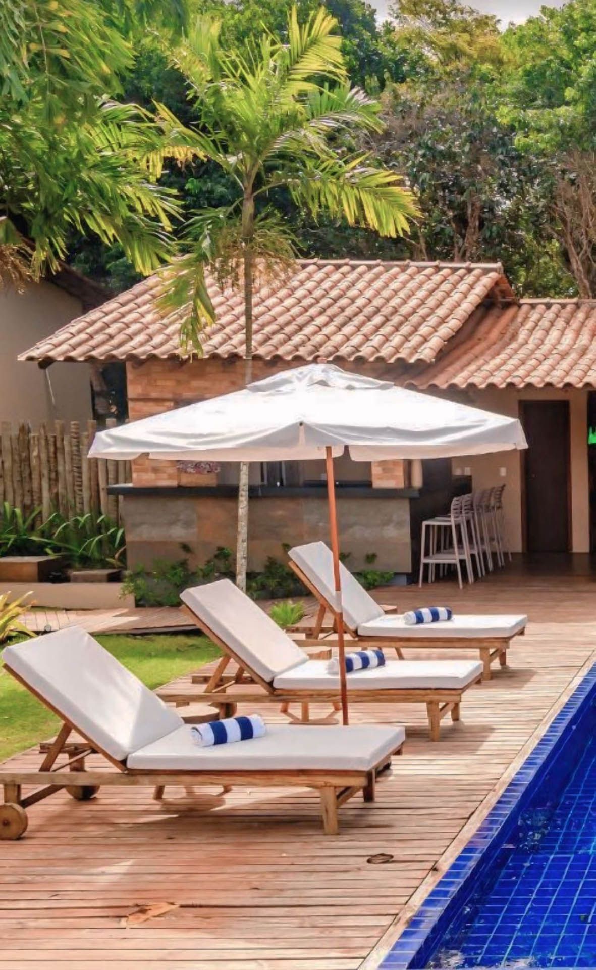 Espreguiçadeiras à beira da piscina sob um guarda-sol branco, ao lado de uma piscina com azulejos azuis e uma cabana tropical.