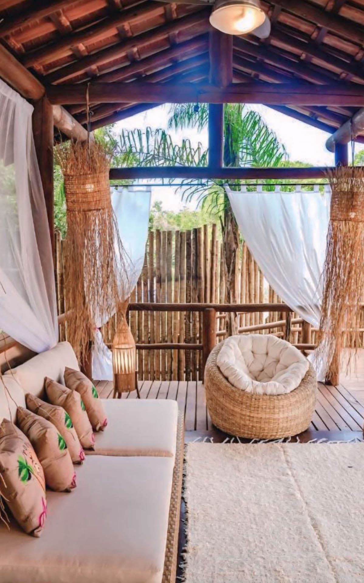 Aconchegante lounge boho com cadeira de vime, sofá e cortinas translúcidas em uma cabana de madeira ensolarada.