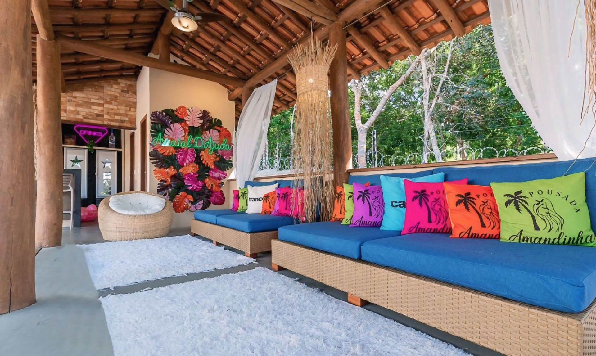 Lounge aconchegante ao ar livre com almofadas azuis, travesseiros coloridos e um teto de palha com vista para a vegetação.