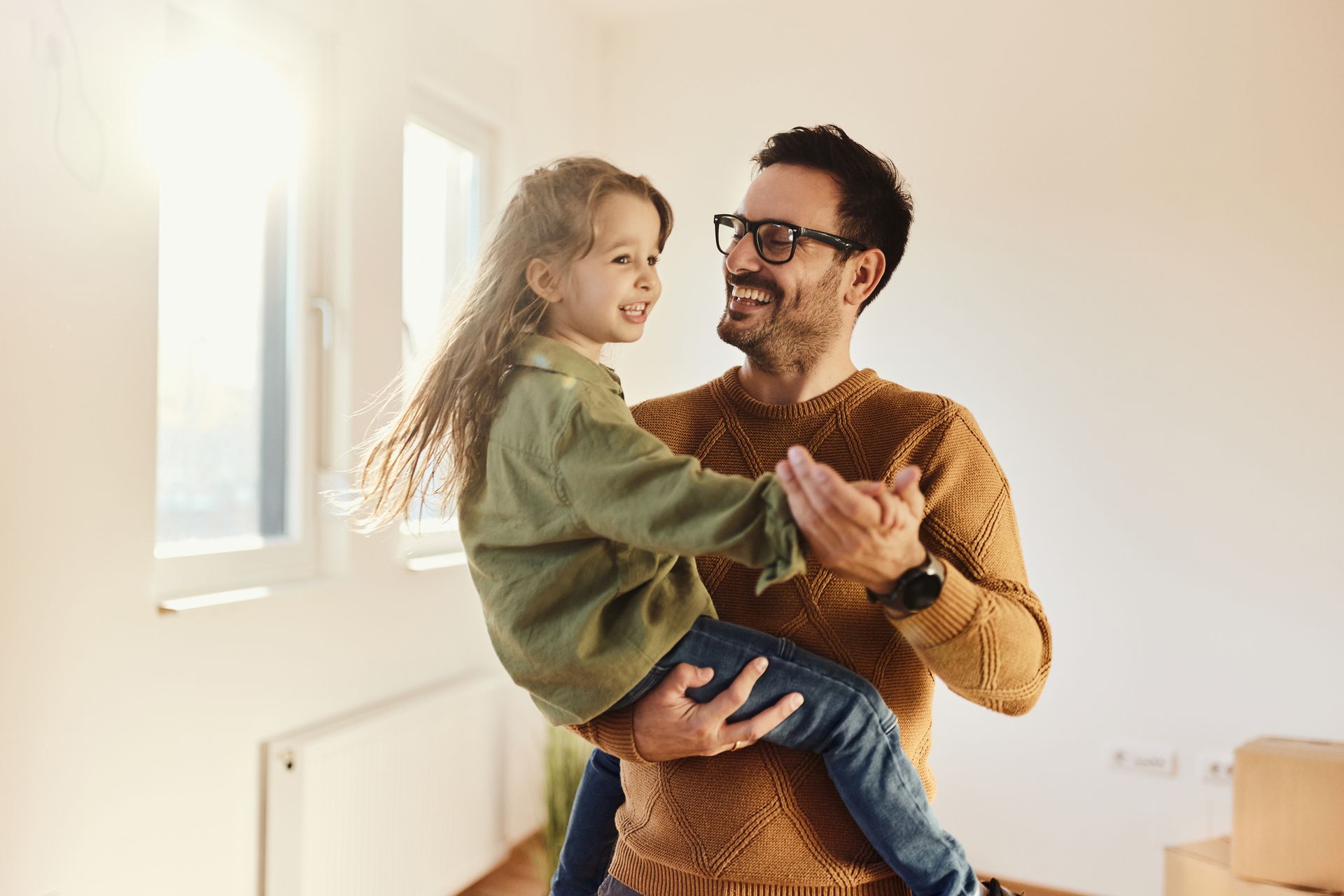 Father in glasses dances with his smiling daughter in a sunny room.