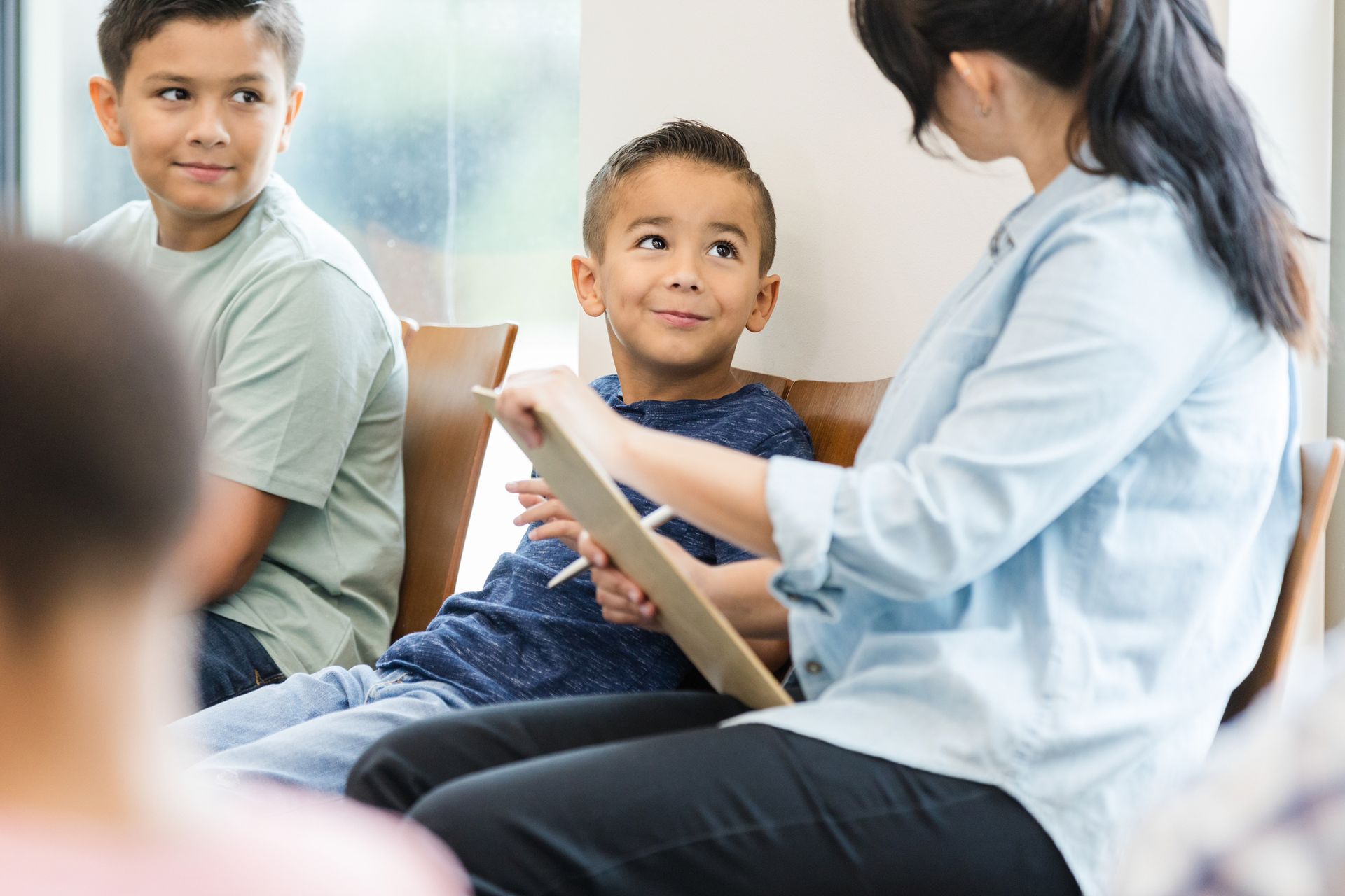 A woman with a clipboard talks to two boys in a circle, smiling; indoor setting.