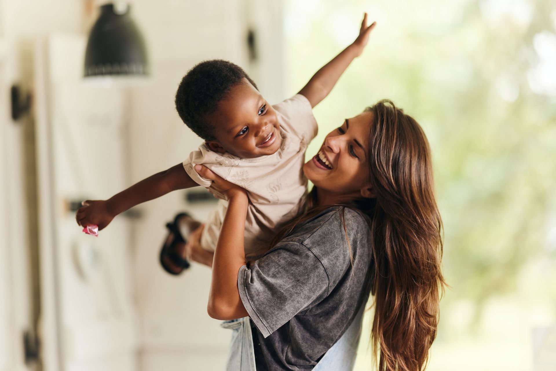 Woman happily holds a child with arms outstretched; both are smiling and laughing indoors.