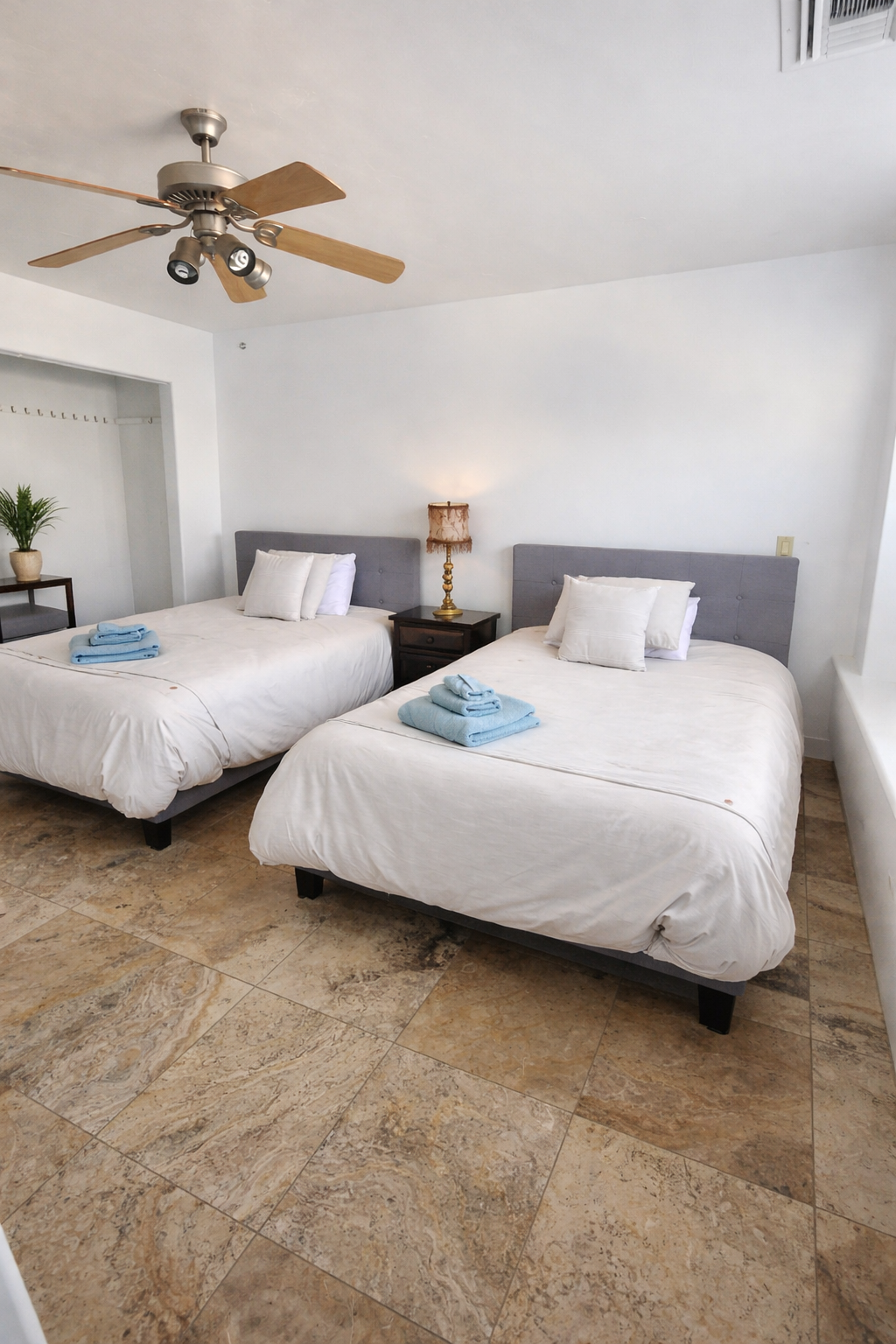 Bright bedroom with two twin beds, white bedding, a nightstand lamp, and a ceiling fan on tiled floors.
