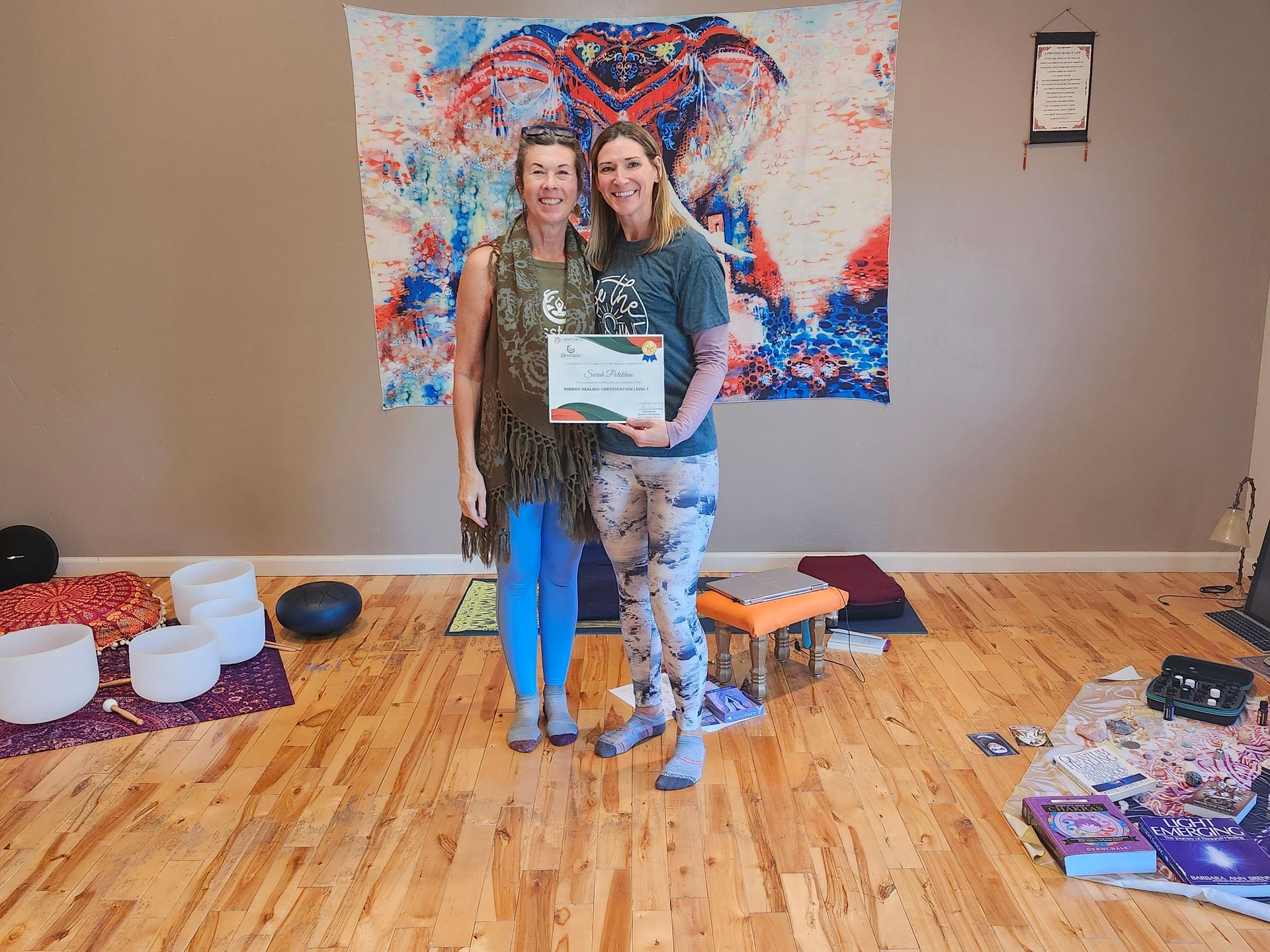 Two women posing with a certificate in front of a colorful heart mural, on a wooden floor stage