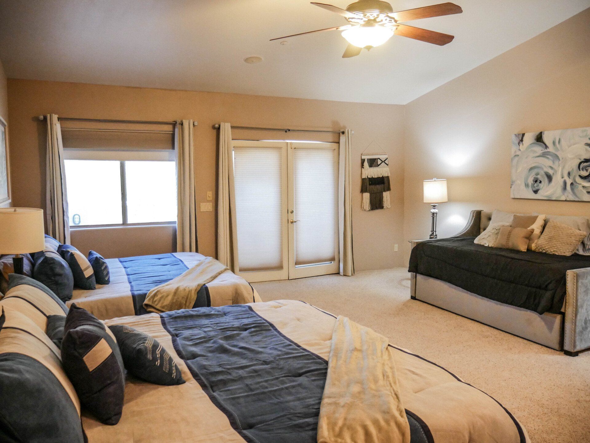 Spacious beige bedroom with two beds, twin windows, ceiling fan, and a dark sofa under wall art