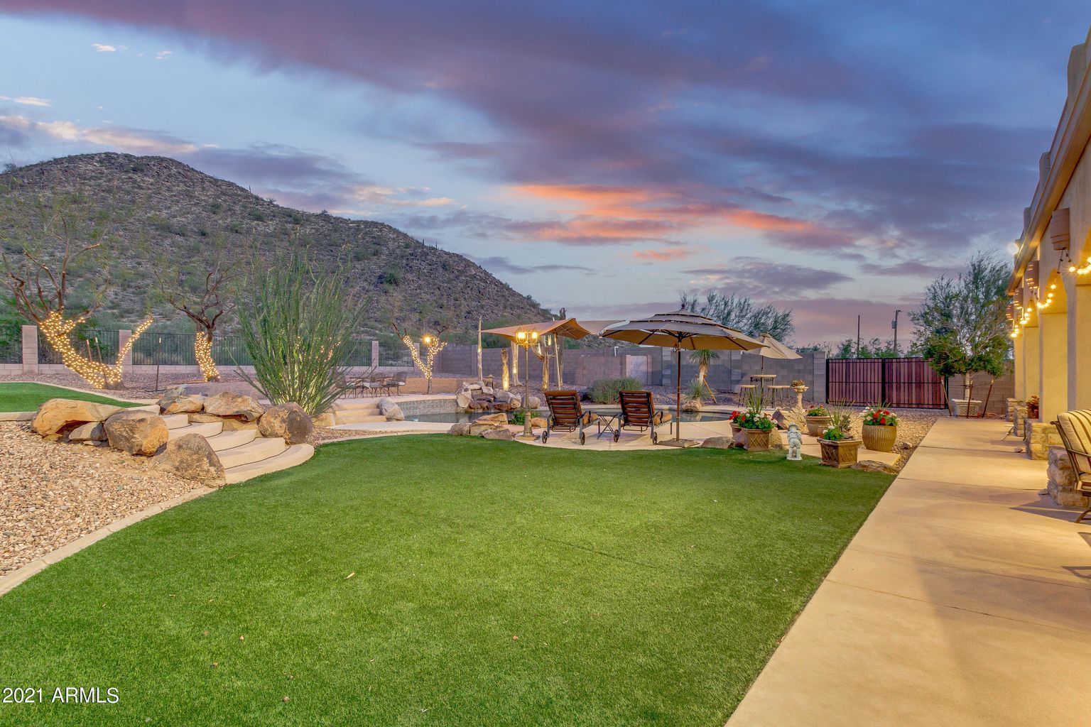 Desert backyard at dusk with green lawn, patio seating, mountain view, and warm string lights