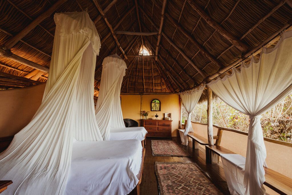 Thatched safari tent bedroom with draped white beds and curtains, warm wood interior, and patterned rug