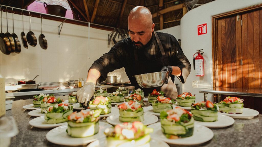 Chef plating multiple salads in a busy kitchen, with dishes lined across a counter.