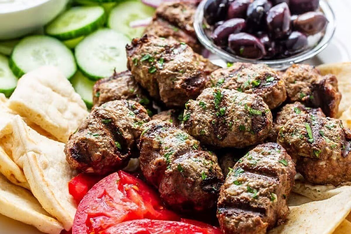 Grilled meat patties with pita, cucumber, olives, and roasted red peppers on a platter