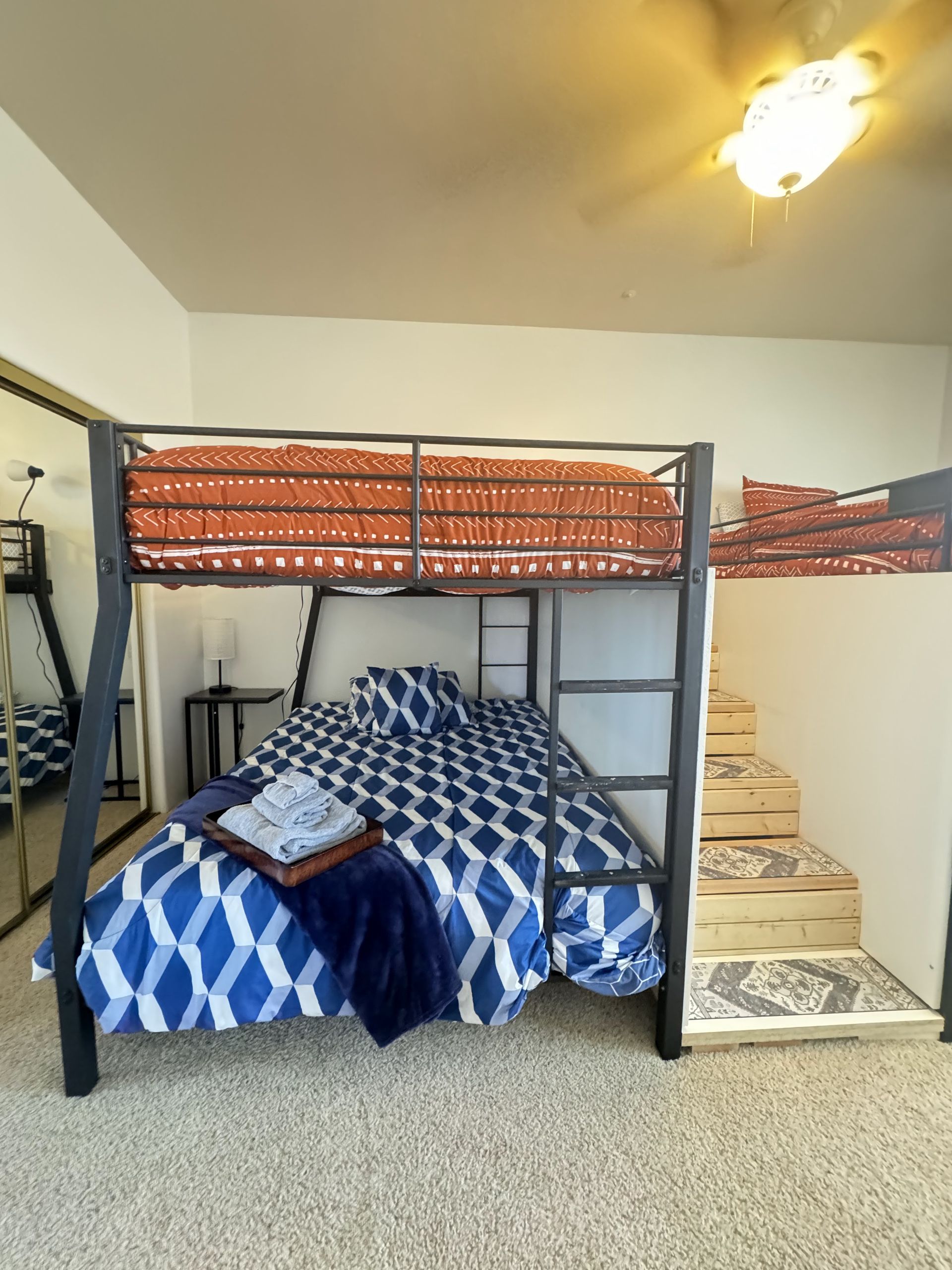 Loft bed with blue patterned bedding in a small bedroom beside stairs and a ceiling fan