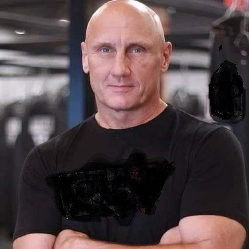 Bald man in a black T-shirt stands with arms crossed in a gym, looking at the camera.