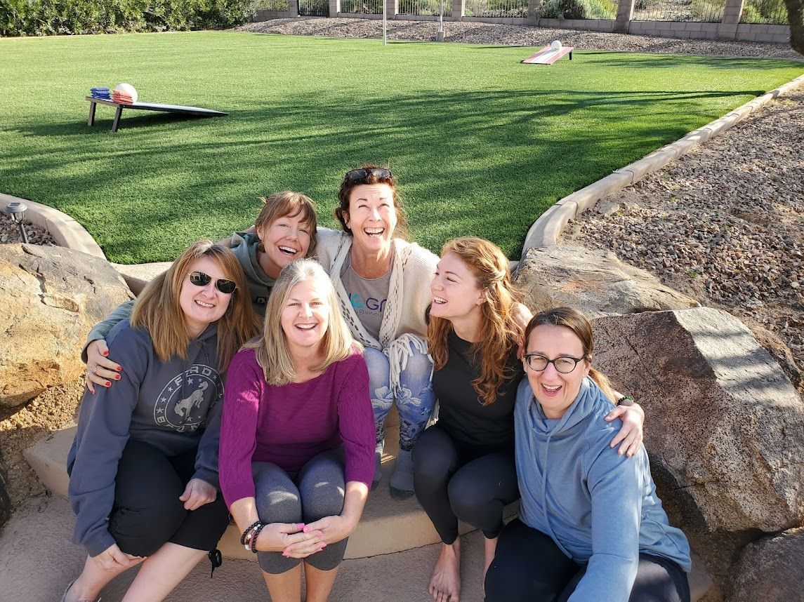 Six people sitting together on a stone bench in a sunny park, smiling at the camera.