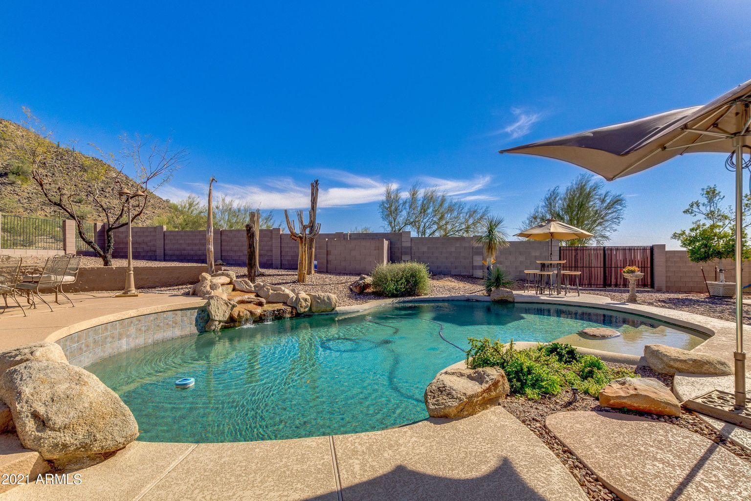 Backyard pool with turquoise water, desert landscaping, rocks, and a shaded patio under a blue sky