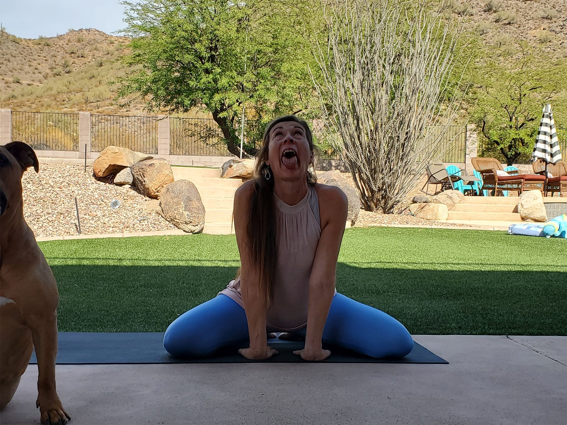 Woman in yoga pose outdoors, mouth open, dog at left.