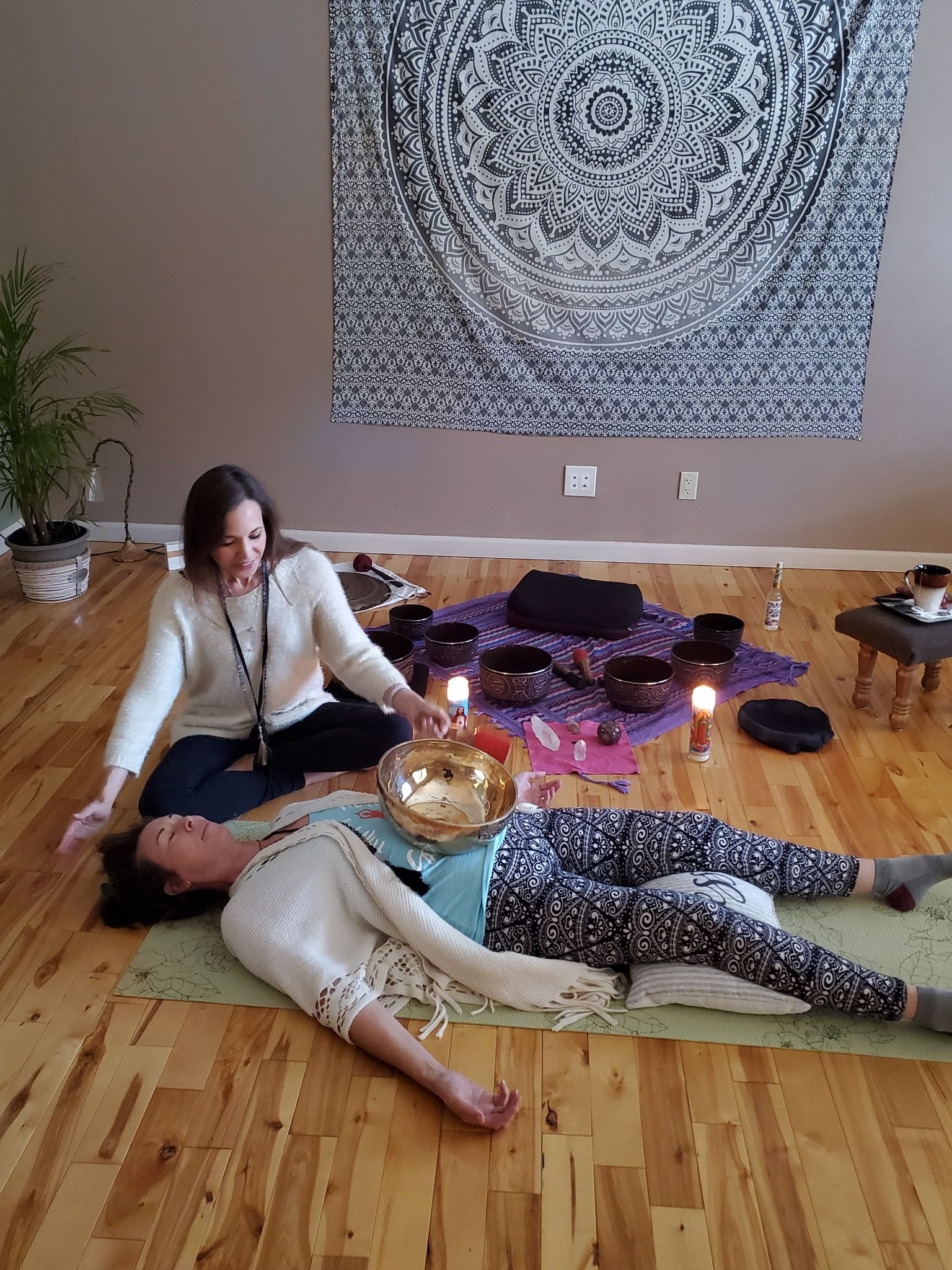Two people lie on yoga mats in a candlelit room with singing bowls and a tapestry on the wall.