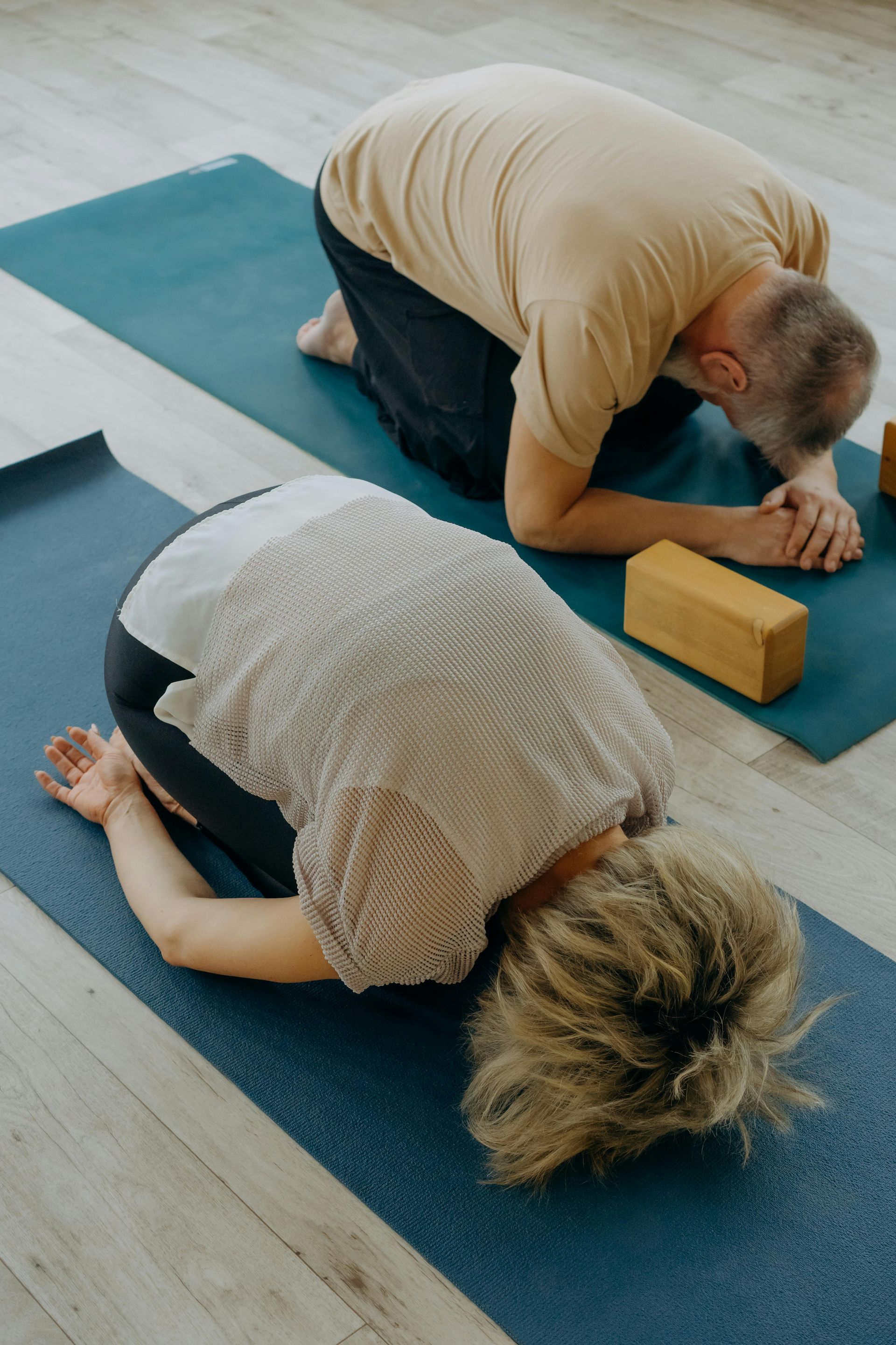 Two people in yoga child’s pose on blue mats, one using a wooden block for support