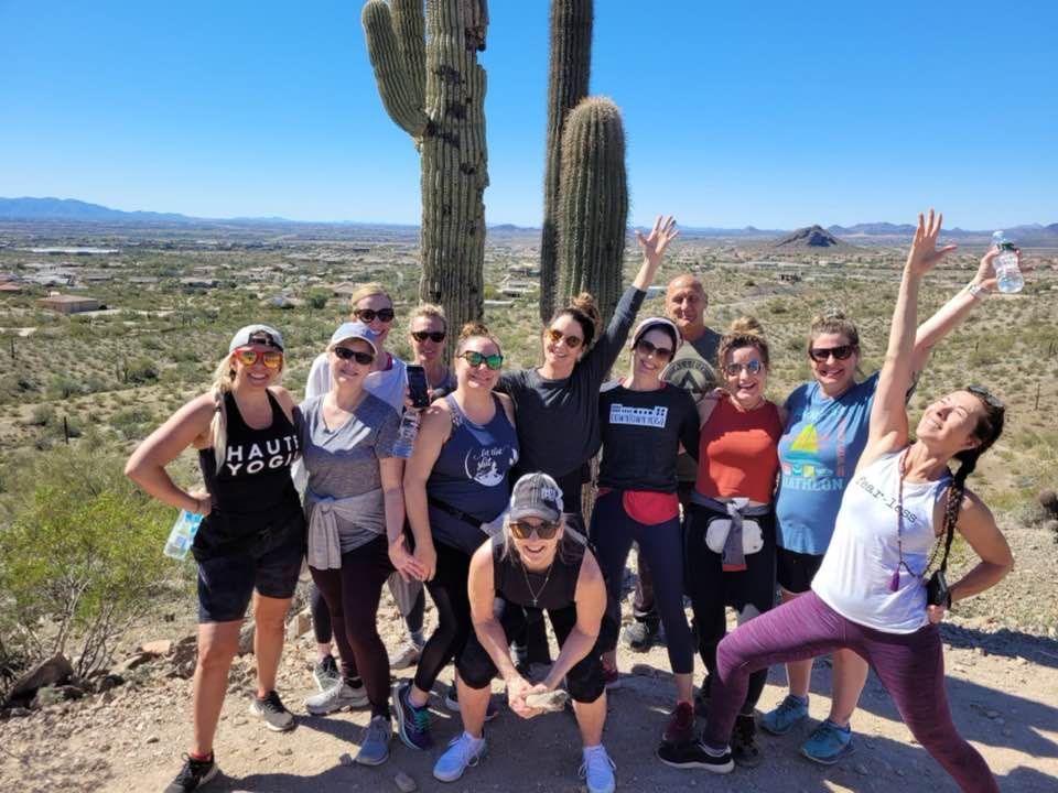 Group of hikers posing by tall cacti in a sunny desert landscape with mountains in the distance