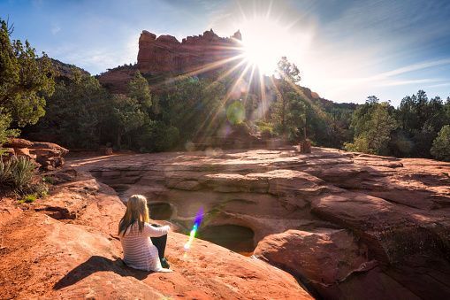 Person sitting by a sunlit desert creek with red rock cliffs and trees in the background