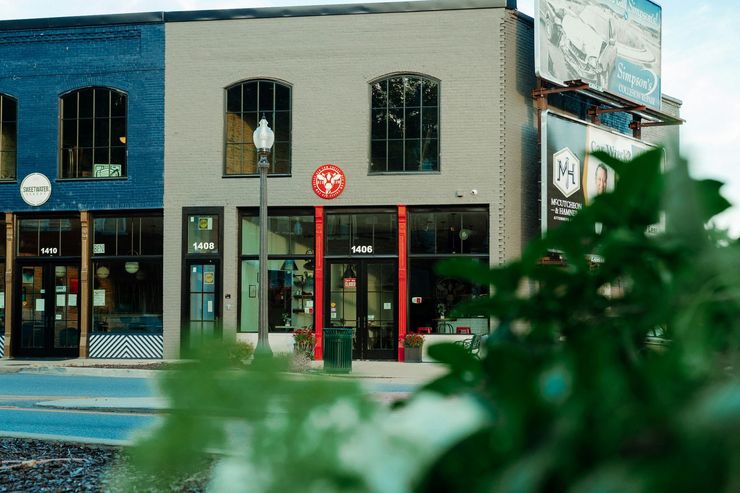 Street view of storefronts with brick facades and a red-framed entrance, seen past blurred green leaves
