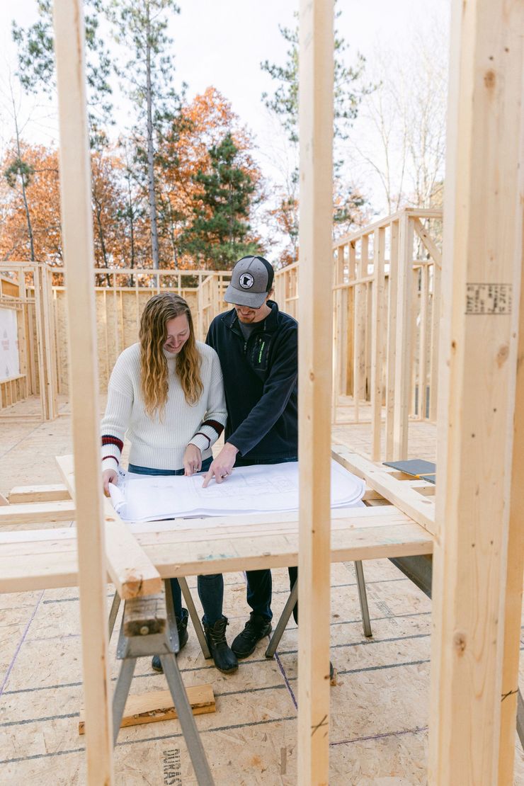 Two people in a wooden frame structure review plans on a table at an outdoor construction site.