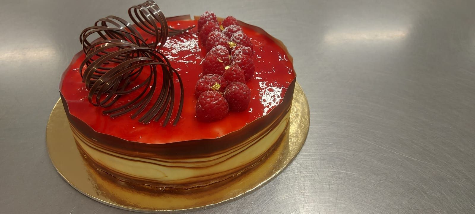 Una torta ai lamponi con glassa rossa, decorazioni di cioccolato e lamponi su un piatto dorato.