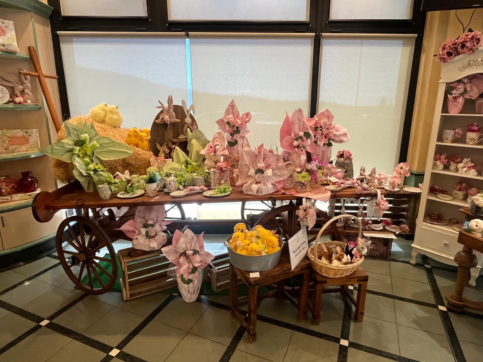 In un negozio è esposto un carrello carico di articoli decorativi, tra cui composizioni floreali rosa e gialle, sullo sfondo di una vetrina.