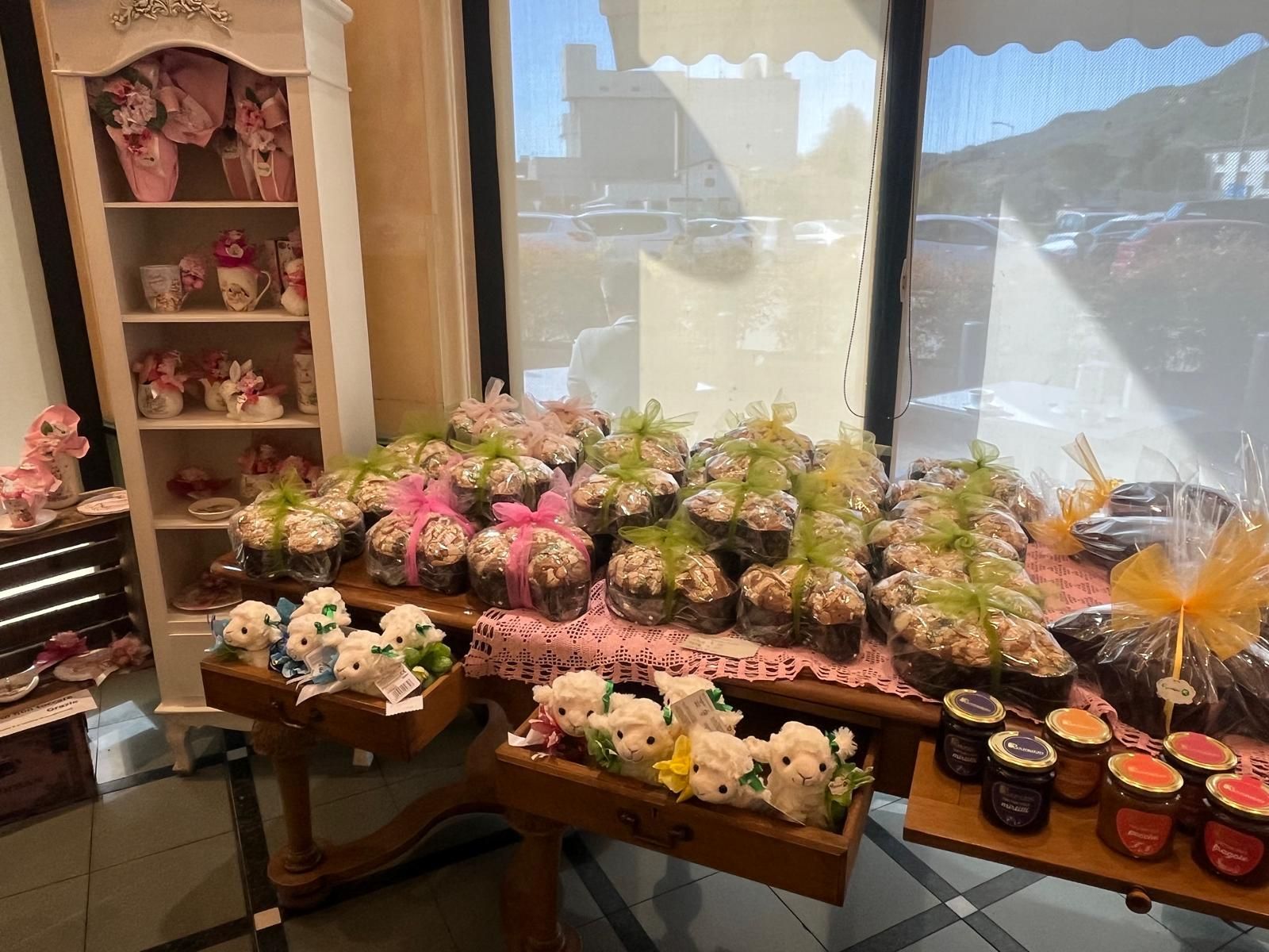 Esposizione di prodotti da forno con dolcetti incartati, marmellate e animali di peluche su tavoli vicino a una finestra.