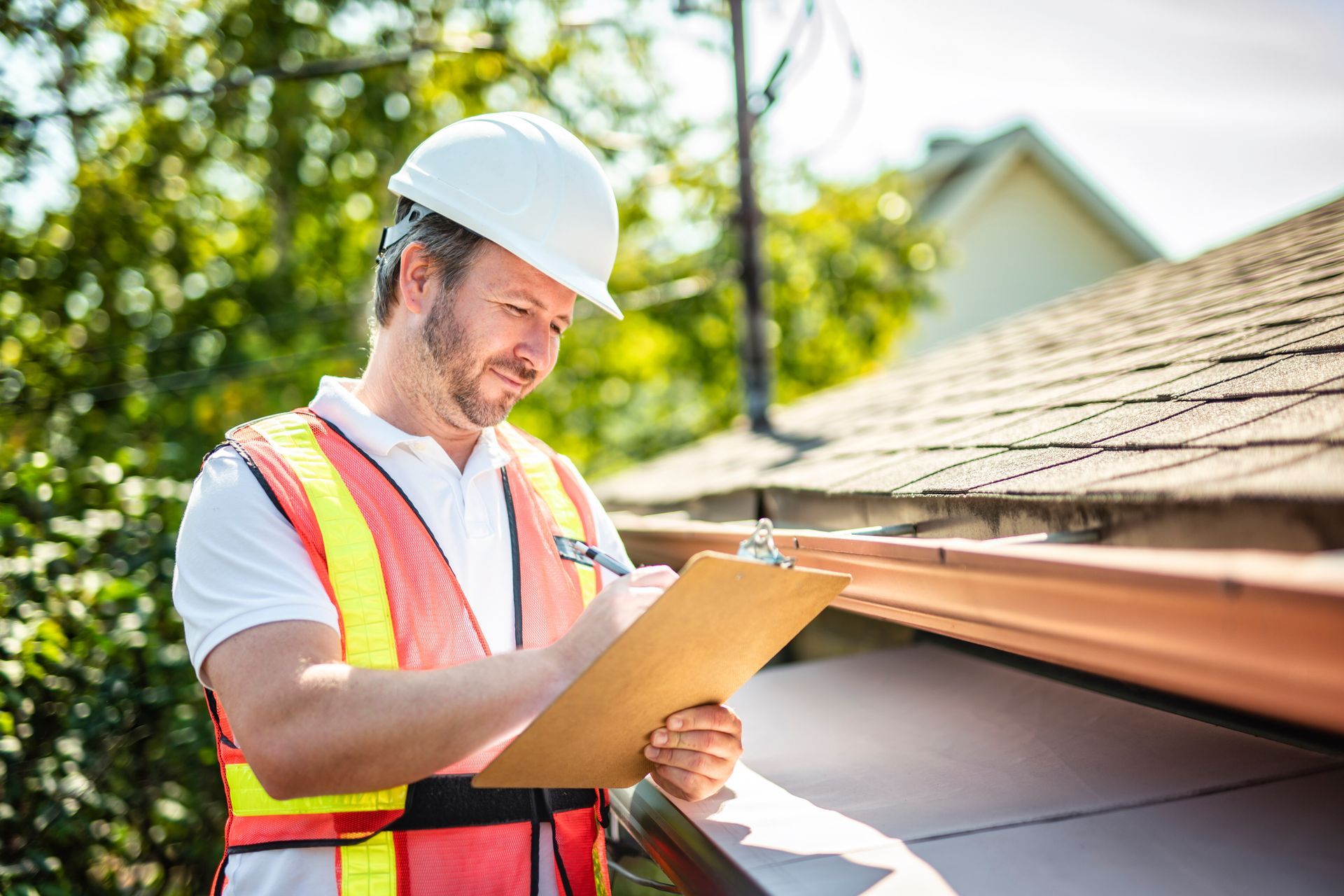 Roofer in hard hat and safety vest inspecting shingle roof edge and gutter, writing on a clipboard. Roofer in hard hat and safety vest inspecting shingle roof edge and gutter, writing on a clipboard.