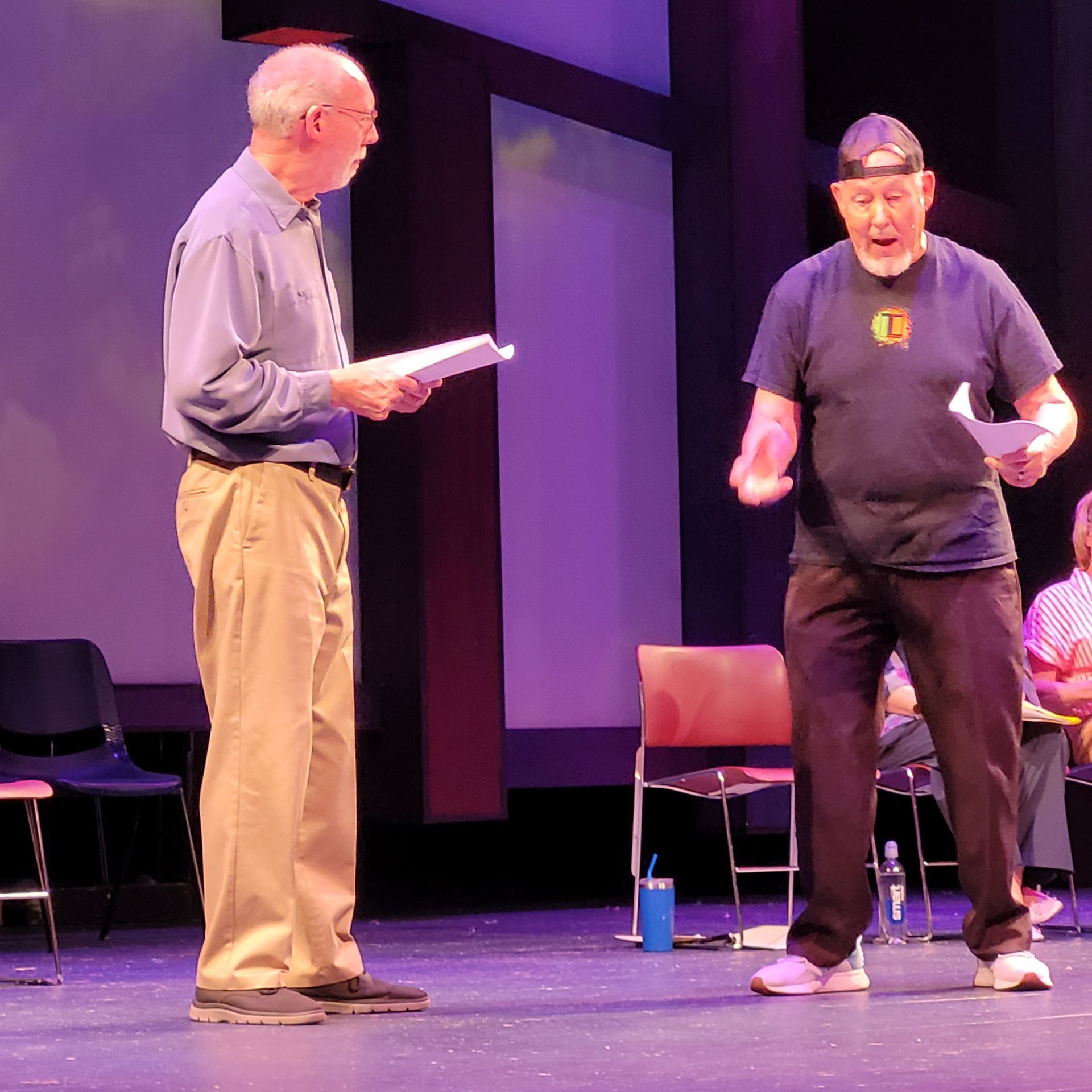 Two men on stage holding scripts, one gesturing, the other looking on. Purple and white backdrop.