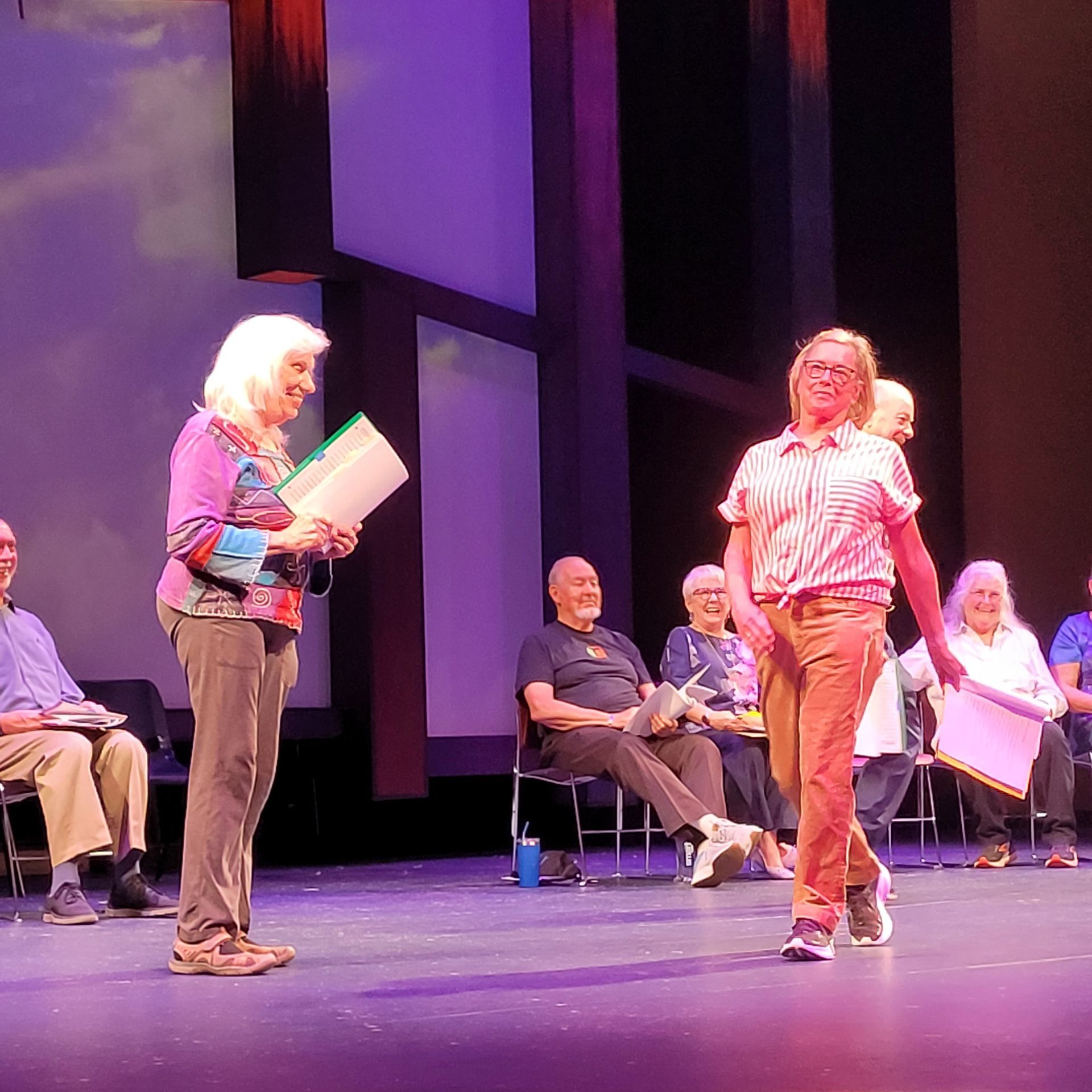 Actors on stage during a play. One woman holds papers, another walks towards her, audience in background.